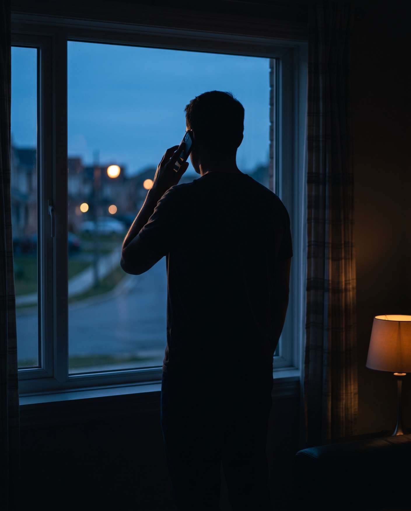 A person standing at a window at dusk with a phone to their ear, the room dark behind them and streetlights coming on outside